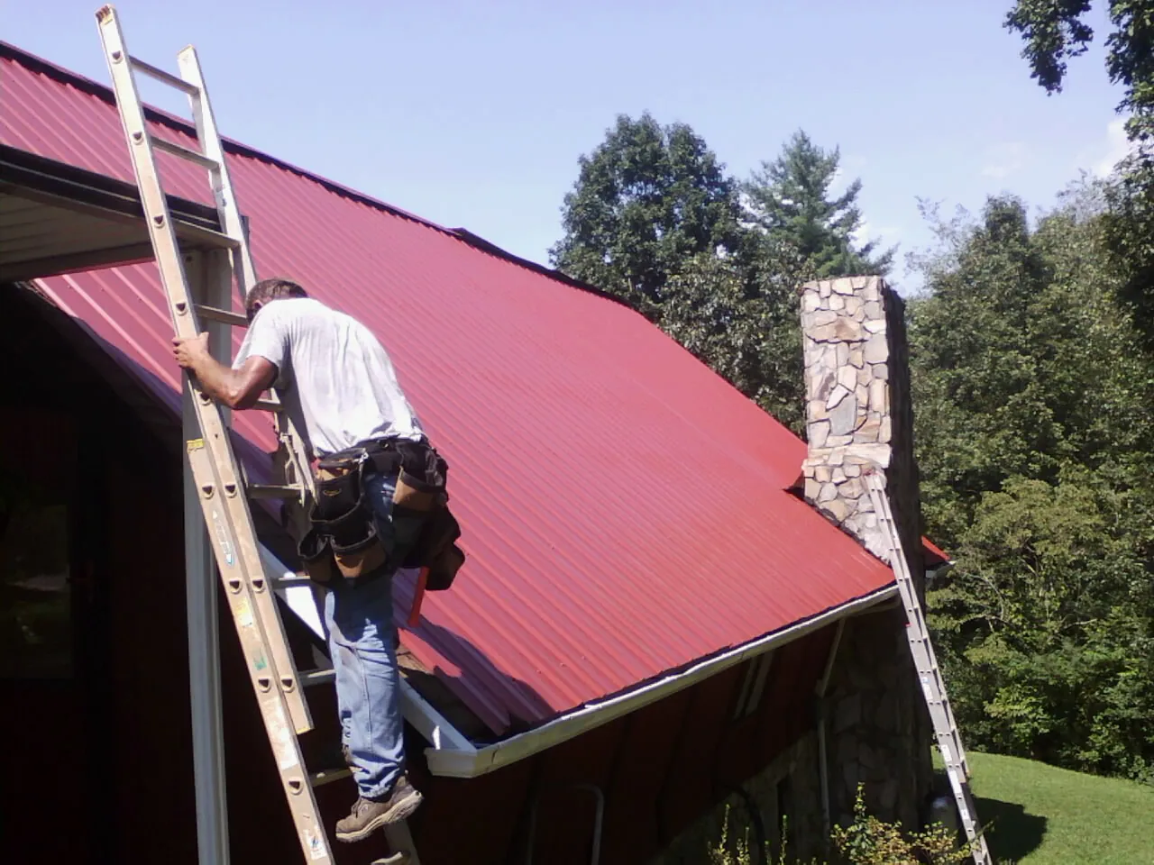 Roofer on a ladder beside a red metal roof and stone chimney — Soffit & Fascia Repair by Roofing Brothers in Brown City